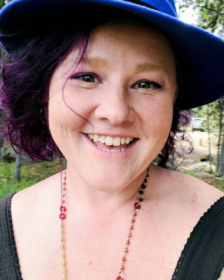 Smiling woman with purple hair, wearing a blue hat and vintage glass necklace.