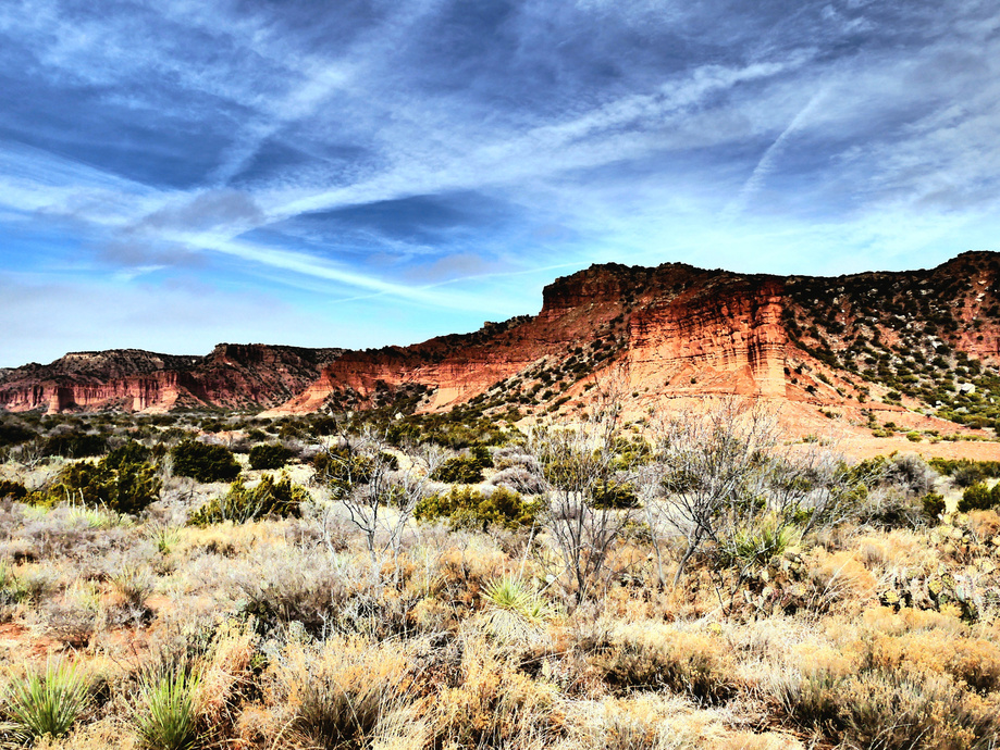 A photo I took and edited of a red-streaked sheer rock formation with green shrubs in the foreground and a bright blue sky with lines of white clouds.