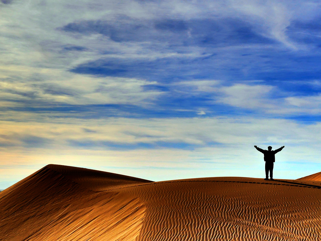 A photo I took and edited showing the silhouette of a figure with their arms raised standing on a waved sand dune with a blue sky and streaky clouds.