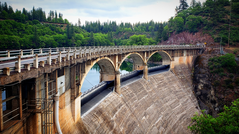 A photo I took and edited of a large concaved, concrete and steel dam with blue water behind it. There are green trees and white clouds.