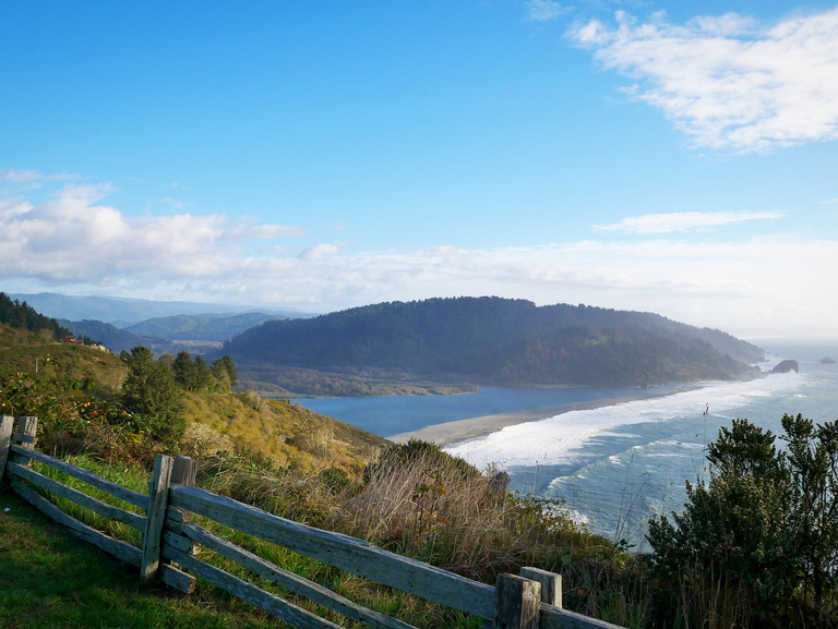 A photo I took of where the Klamath river meets the Pacific ocean. Blue sky with rolling green hills and a long white cap wave braking along the sand bar.